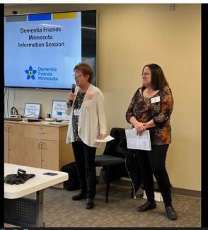 Two people standing at the front of a room. Screen with Dementia Friends information visible.